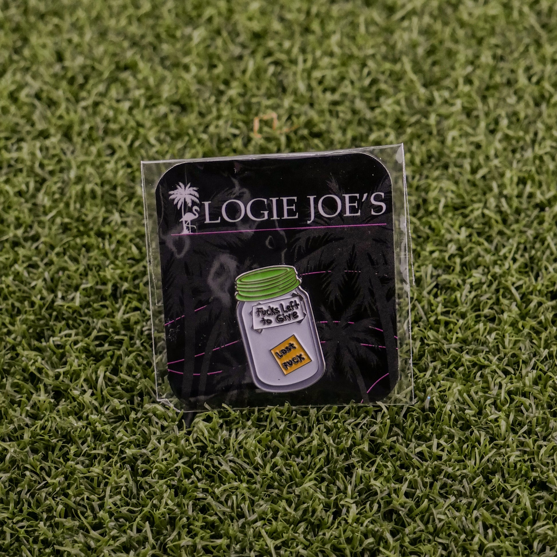 The Jar of “F”s enamel pin by Ballmarkers by Logie Joes sits on a branded card atop artificial grass—perfect as a funny ball marker or gift for fans of unique golf accessories.