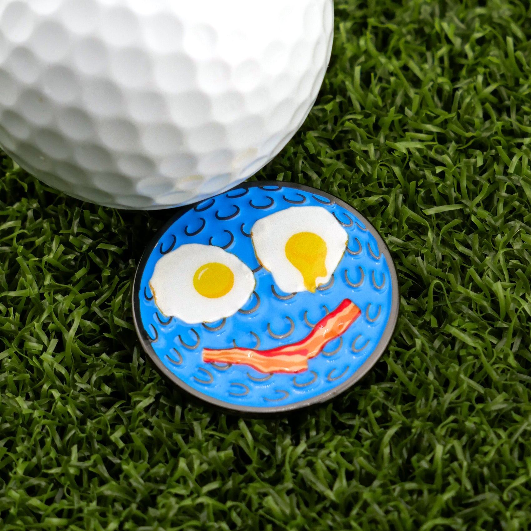 The Breakfast Ball by Logie Joe's Golf is a blue ball marker with a cartoon face—large white eyes and a red smile—adding fun to your golf gear as it sits on the green next to a white golf ball.