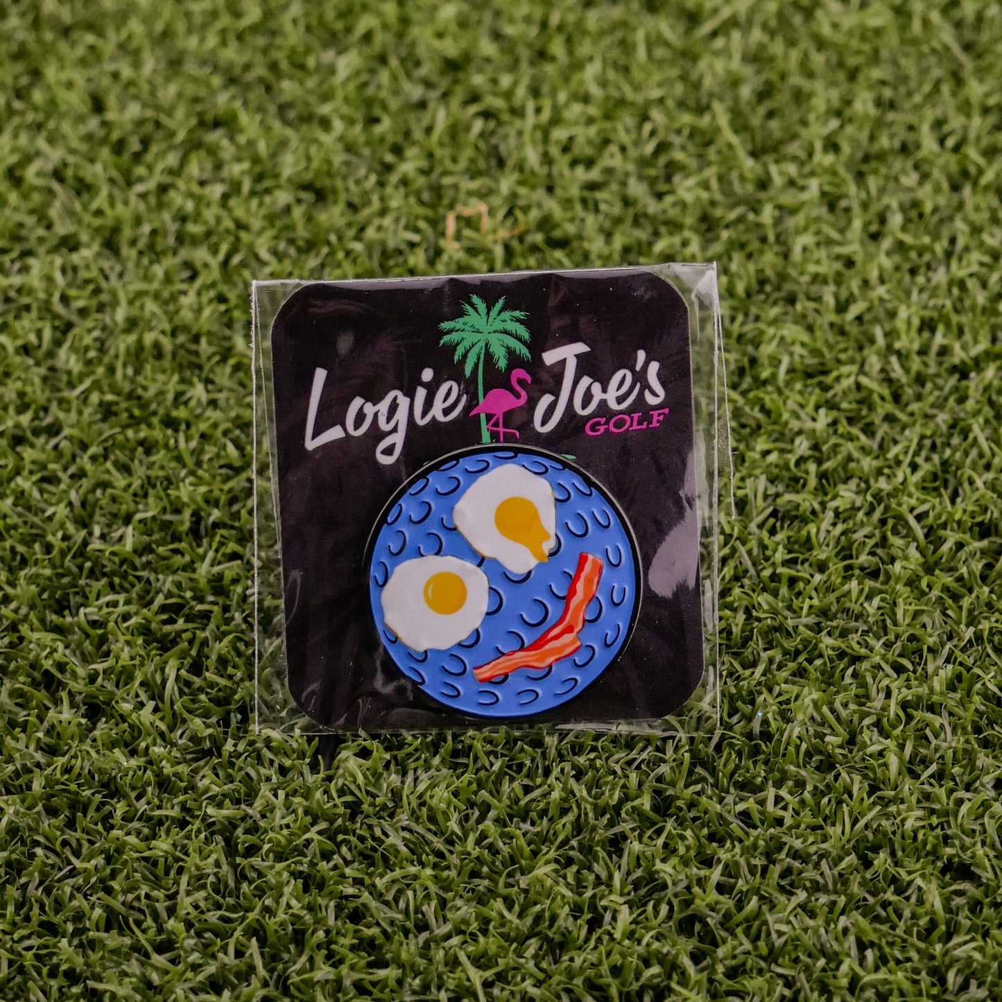 The Breakfast Ball pin by Logie Joe's Golf, shaped like a golf ball with fried egg eyes and a bacon mouth, comes on grass with branded card—making it an ideal premium ball marker.