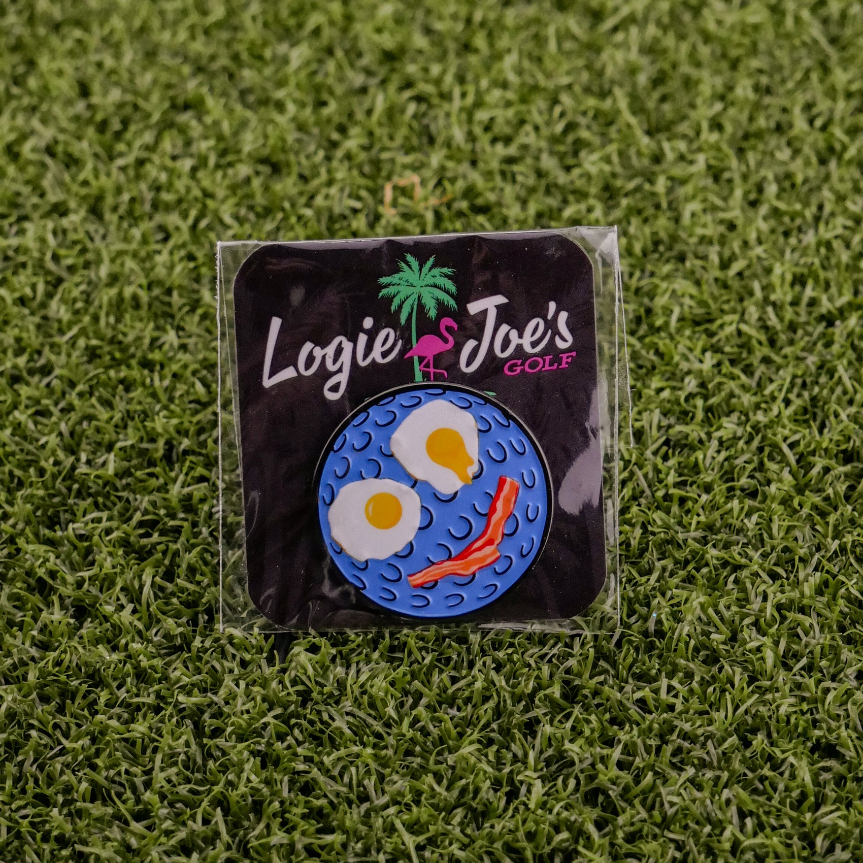 The Breakfast Ball pin by Logie Joe's Golf, shaped like a golf ball with fried egg eyes and a bacon mouth, comes on grass with branded card—making it an ideal premium ball marker.