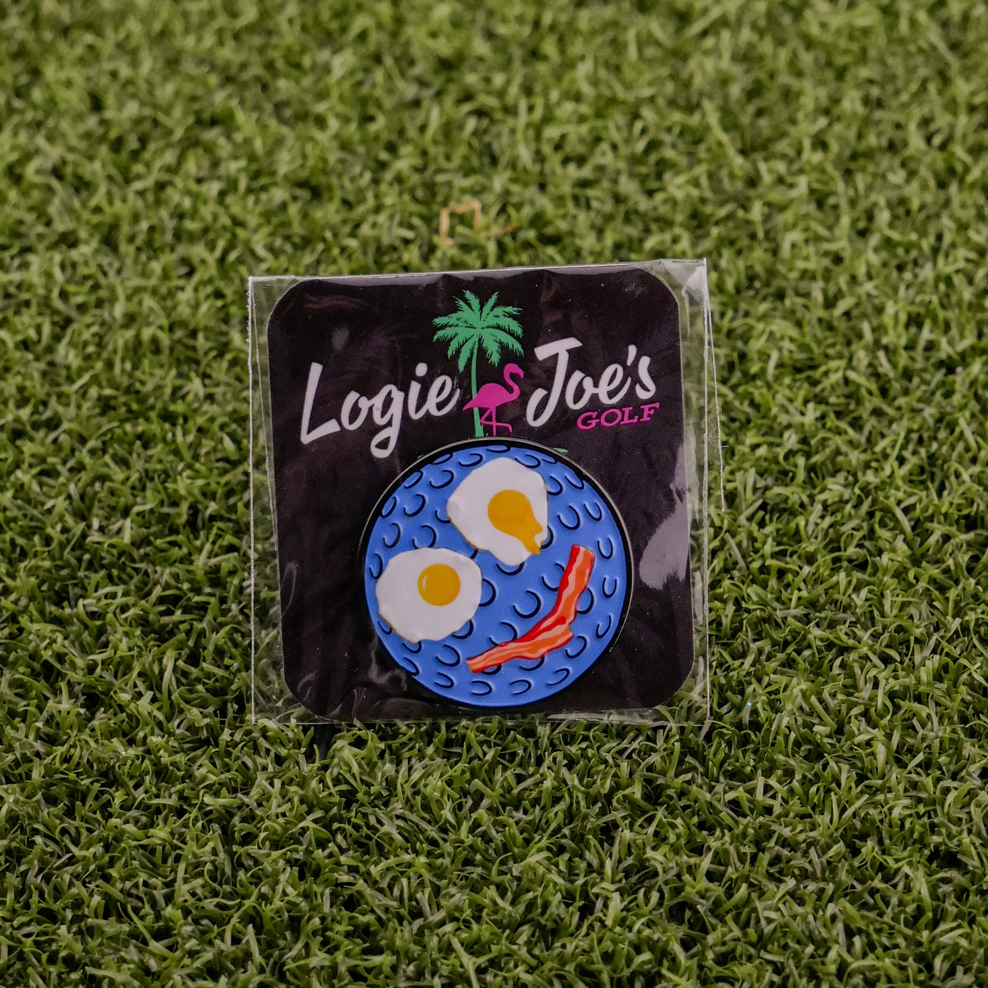 The Breakfast Ball pin by Logie Joe's Golf, shaped like a golf ball with fried egg eyes and a bacon mouth, comes on grass with branded card—making it an ideal premium ball marker.