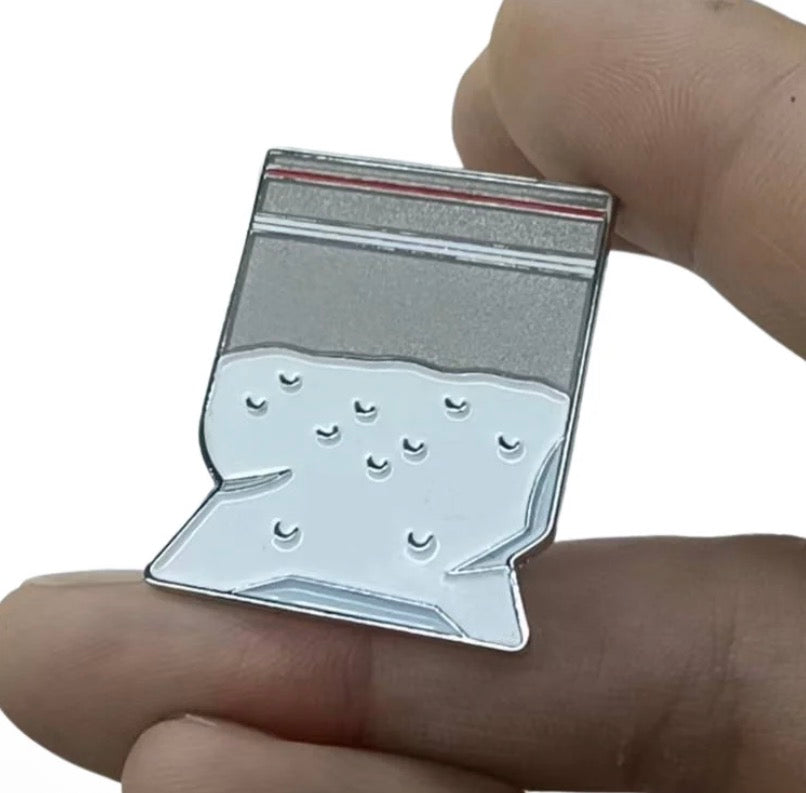 A hand holds the Tee Bumps Ground Score Ball Marker, an enamel pin designed to resemble a small plastic bag filled with white powder and dots—an eye-catching accessory that’s perfect for collectors or as a quirky golf ball marker for those with a sense of humor.