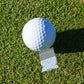 A golf ball rests on green grass beside a Ground Score Ball Marker by Tee Bumps—a small, clear plastic bag containing white granules that doubles as a unique marker and casts a subtle shadow during your round.
