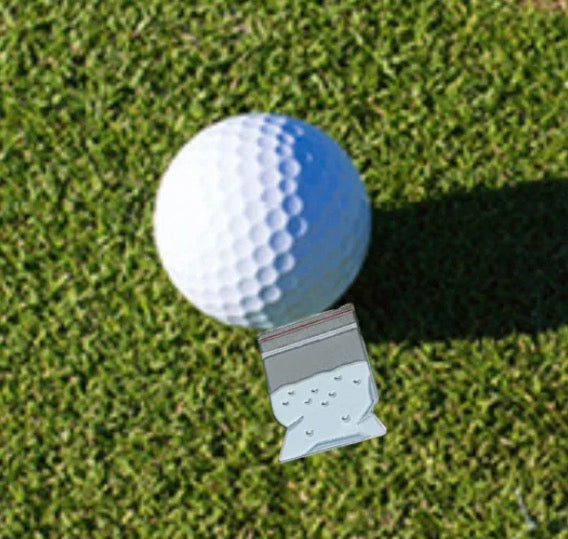 A golf ball rests on green grass beside a Ground Score Ball Marker by Tee Bumps—a small, clear plastic bag containing white granules that doubles as a unique marker and casts a subtle shadow during your round.