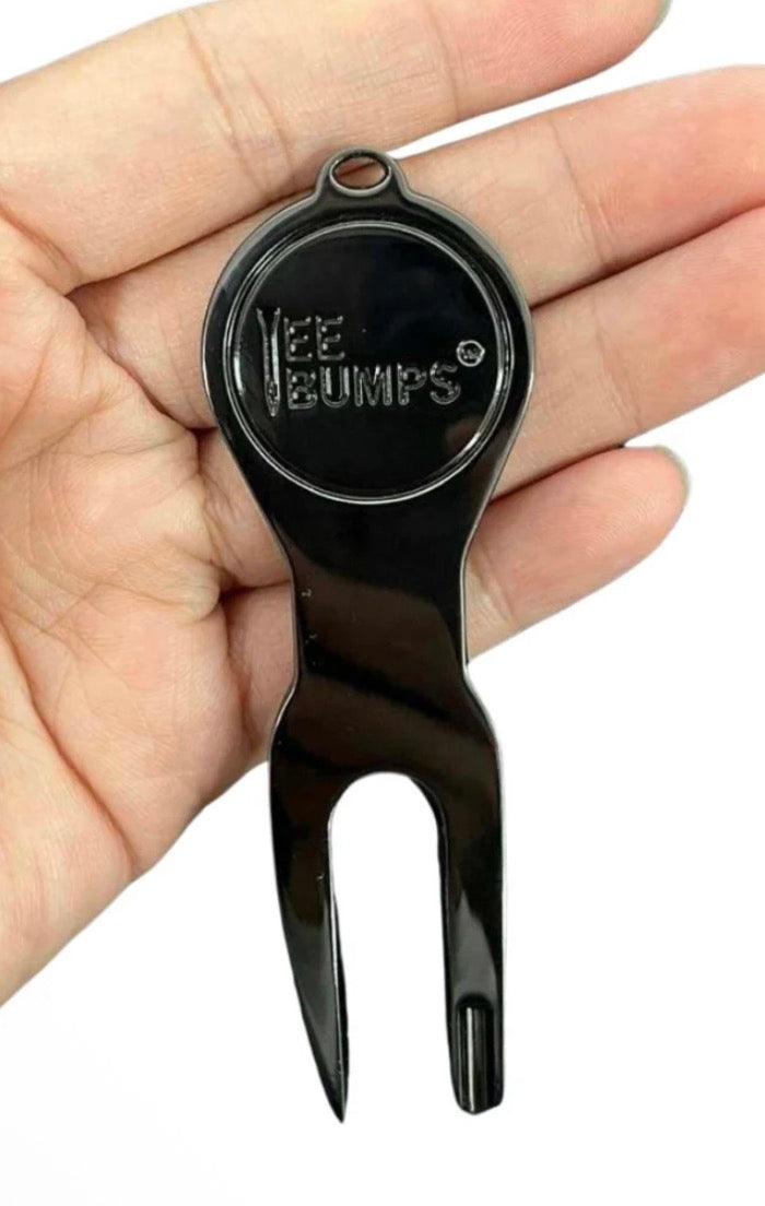 A hand holding “The Deke” Divot Tool by Tee Bumps—a black plastic tool featuring a circular top and pronged fork-like end—an ideal accessory for any golf outing.
