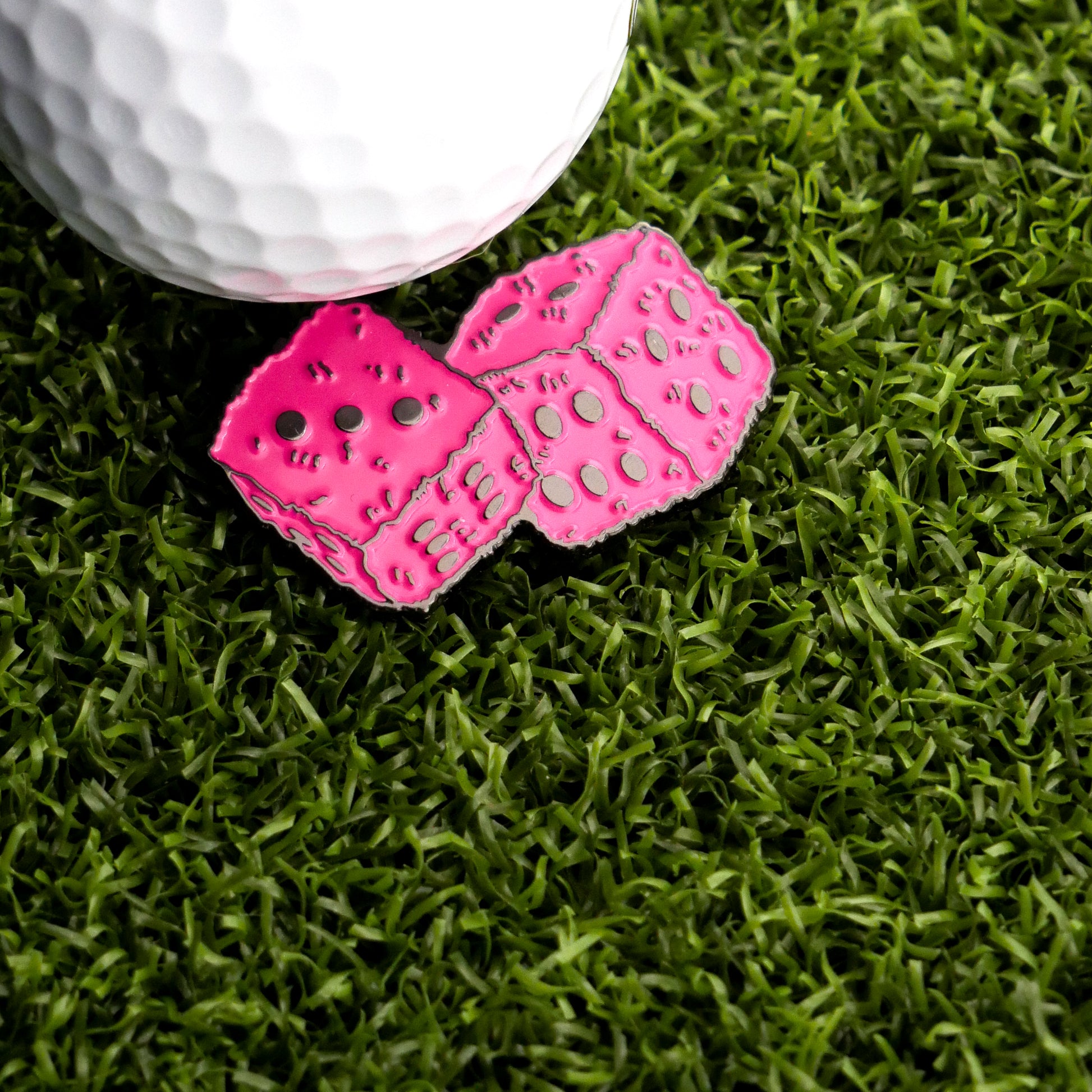 The Pink Fuzzy Dice Ball Marker by Ballmarkers.com rests on green artificial grass beside a white golf marker.