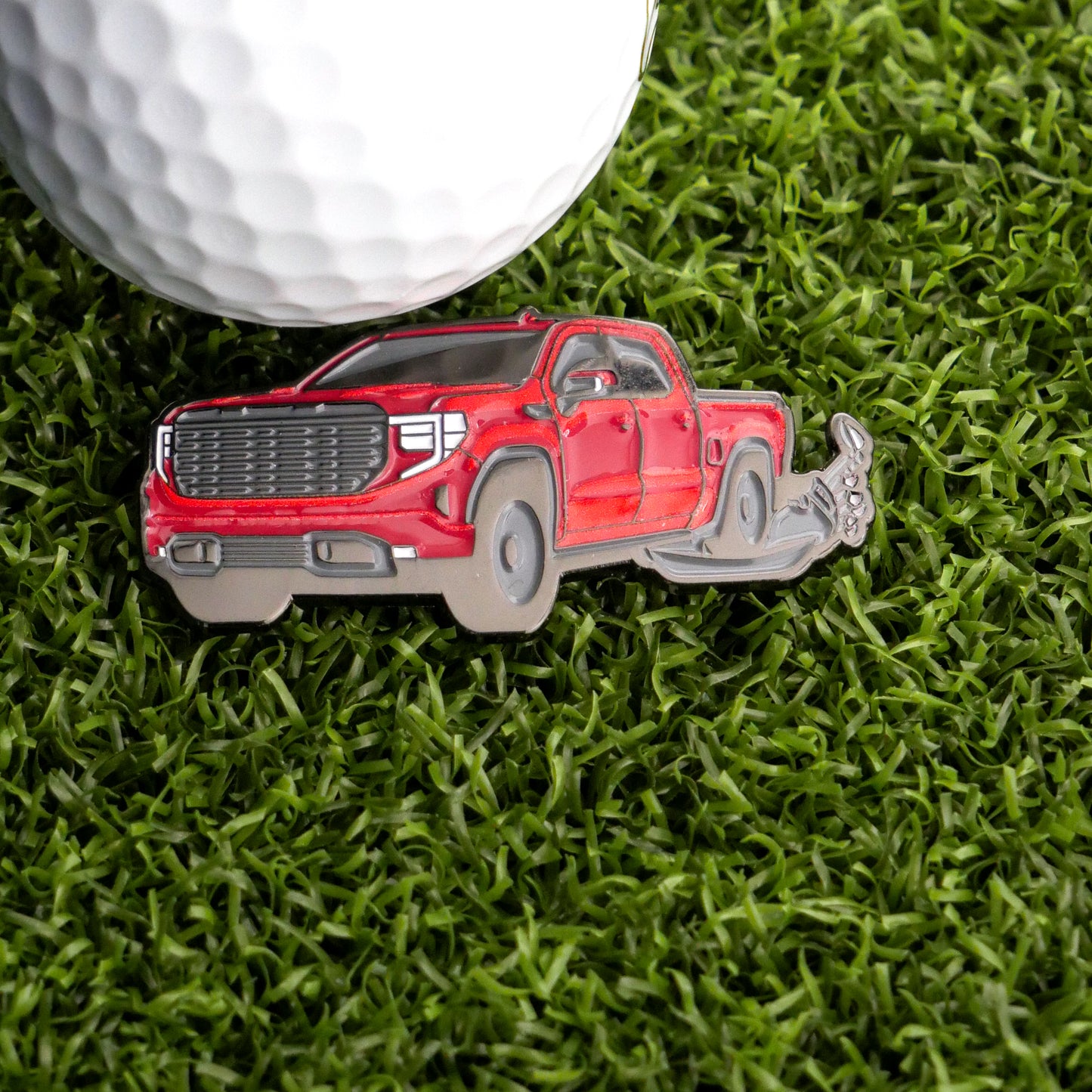 The Logie Joe's Golf Truck Running Over Clubs Ball Marker is a playful pin shaped like a red pickup truck, shown on green grass beside a white golf ball—perfect for expressing some on-course frustration.