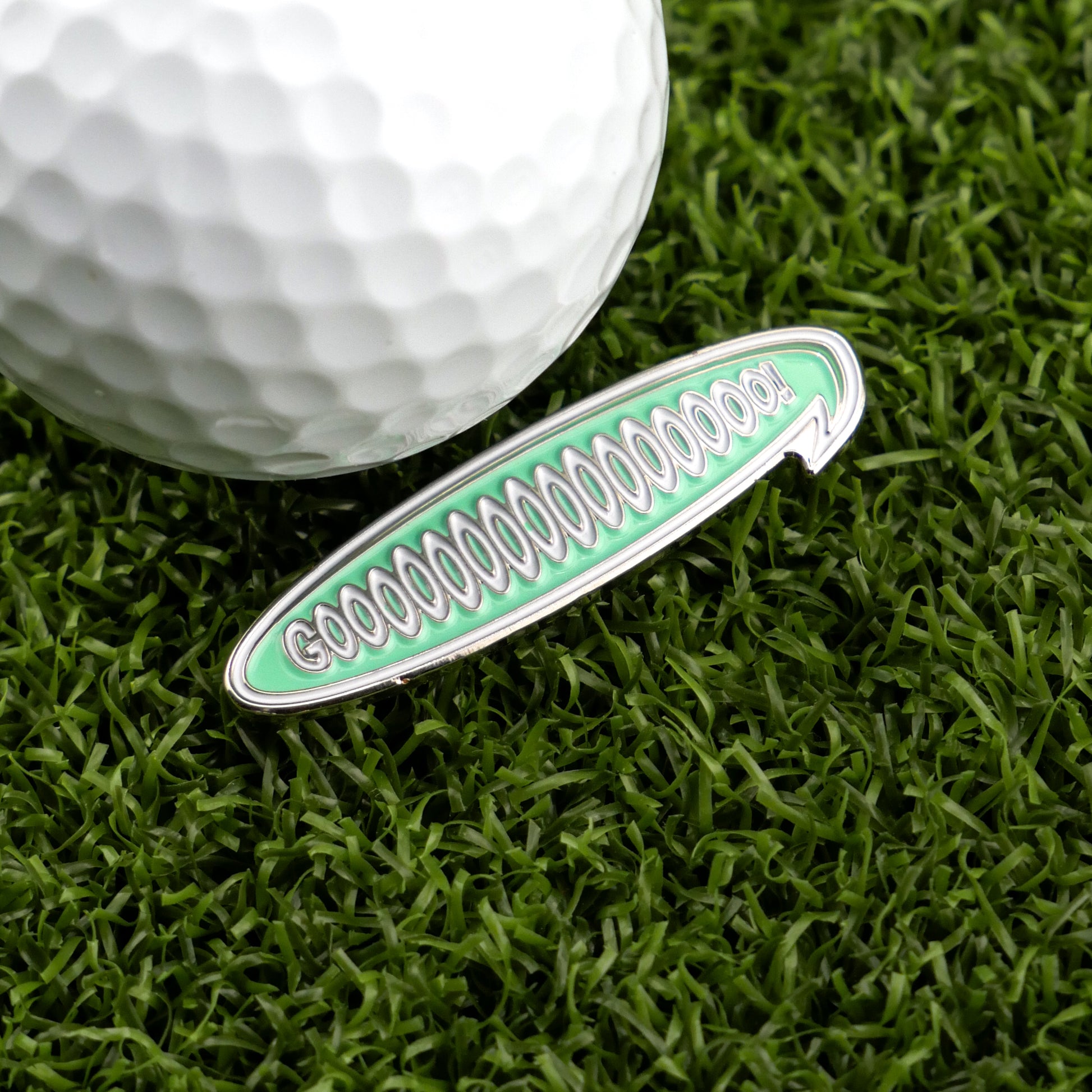 A white golf ball and Logie Joe's Golf Gooooooooooo! Speech Bubble marker in green with silver lettering rest on artificial grass—ideal for fans of unique, personalized golf accessories.