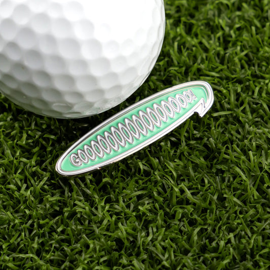 A close-up of a golf ball and the green oval Gooooooooooo! Speech Bubble Ball Marker by Logie Joe's Golf on artificial grass—this playful accessory is perfect for adding cheering spirit to your game as a colorful golf ball marker.