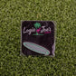 A square black golf pouch from Logie Joe's Golf with a pink flamingo logo rests on green turf, holding custom accessories. Beside it is a "Gooooooooooo! Speech Bubble" pin, both perfect for golf fans.