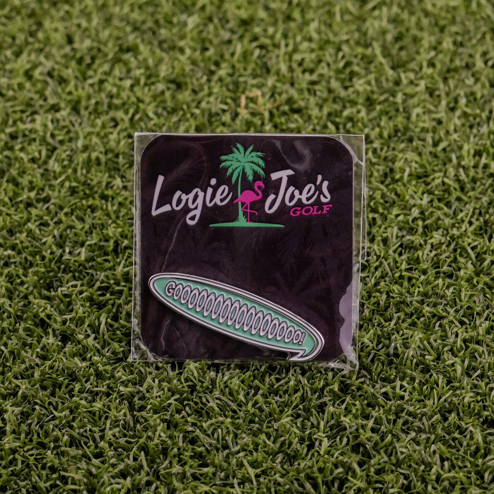 A square black golf pouch from Logie Joe's Golf with a pink flamingo logo rests on green turf, holding custom accessories. Beside it is a "Gooooooooooo! Speech Bubble" pin, both perfect for golf fans.