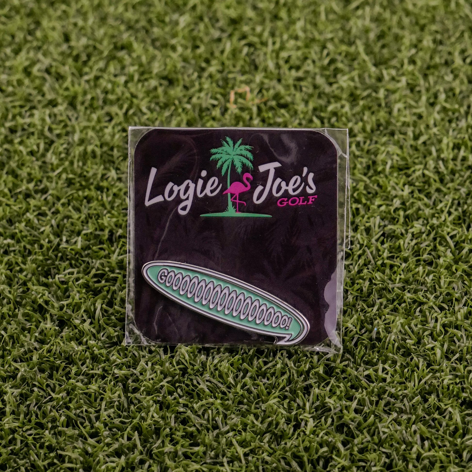 A square black golf pouch from Logie Joe's Golf with a pink flamingo logo rests on green turf, holding custom accessories. Beside it is a "Gooooooooooo! Speech Bubble" pin, both perfect for golf fans.