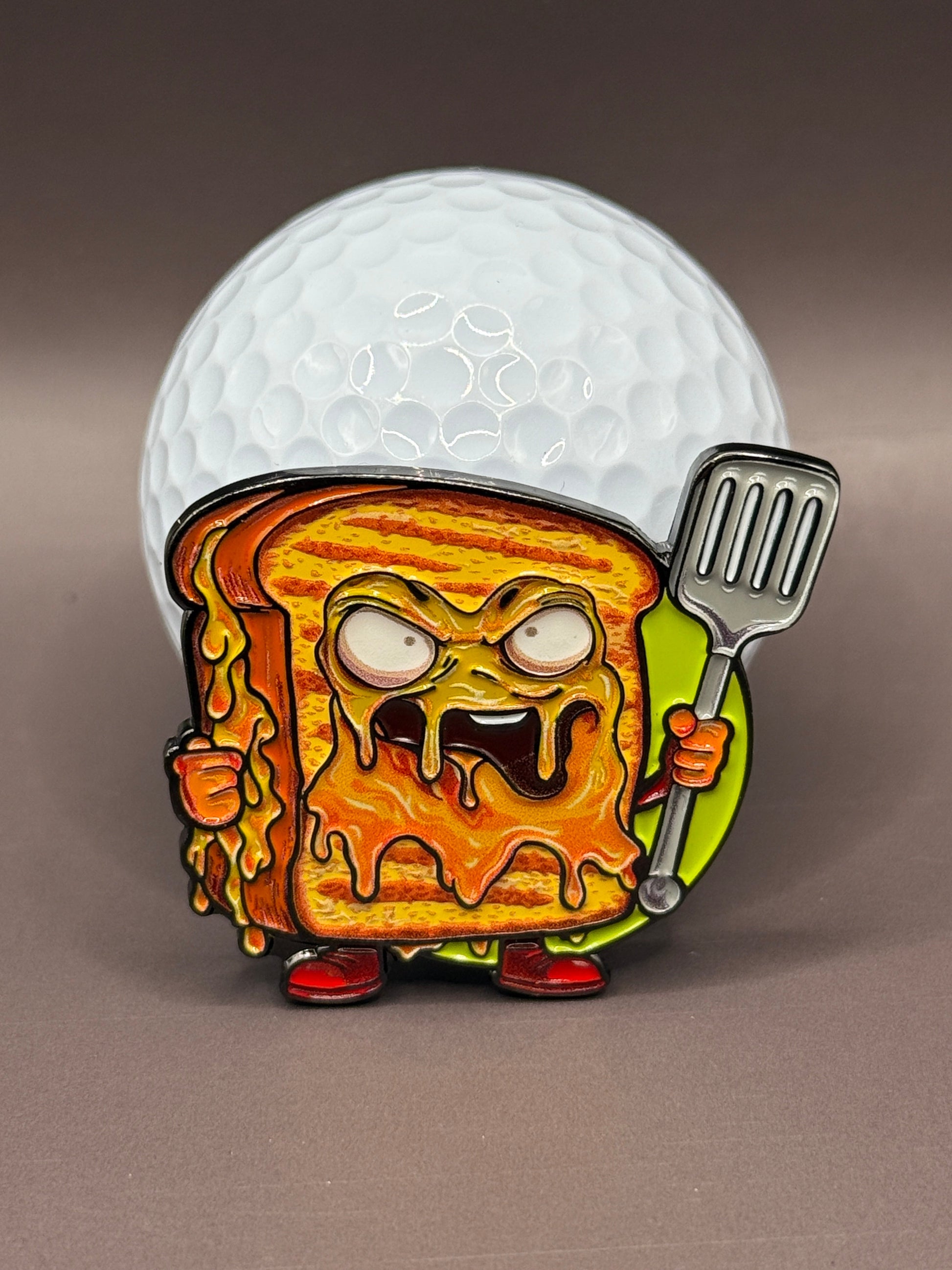 The Gill Cheese Ball Marker by Crown Golf Designs features a cartoonishly angry grilled cheese character with a metal spatula, standing before a giant golf ball, with melted cheese dripping and a fierce look.