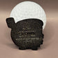 A white golf ball stands behind a black, metal Gill Cheese Ball Marker by Crown Golf Designs, featuring "CROWN GOLF PIGGIE SERIES 0142" and a crown symbol, set against a plain brown background.