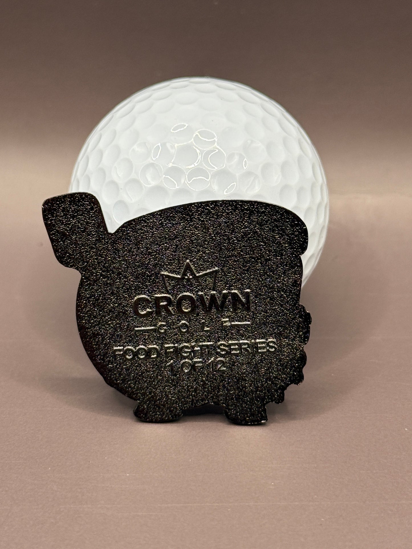 A white golf ball stands behind a black, metal Gill Cheese Ball Marker by Crown Golf Designs, featuring "CROWN GOLF PIGGIE SERIES 0142" and a crown symbol, set against a plain brown background.