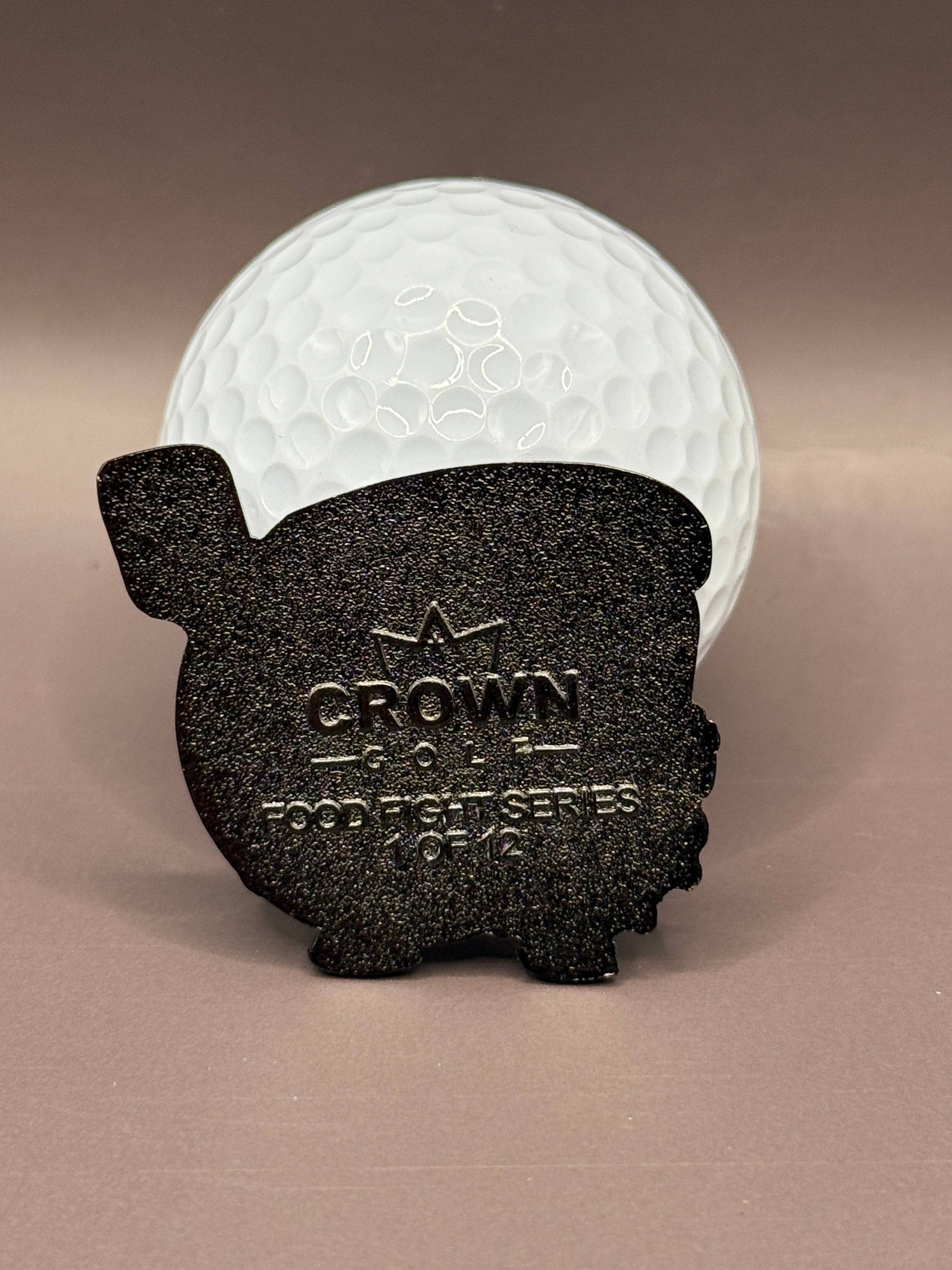 A white golf ball stands behind a black, metal Gill Cheese Ball Marker by Crown Golf Designs, featuring "CROWN GOLF PIGGIE SERIES 0142" and a crown symbol, set against a plain brown background.