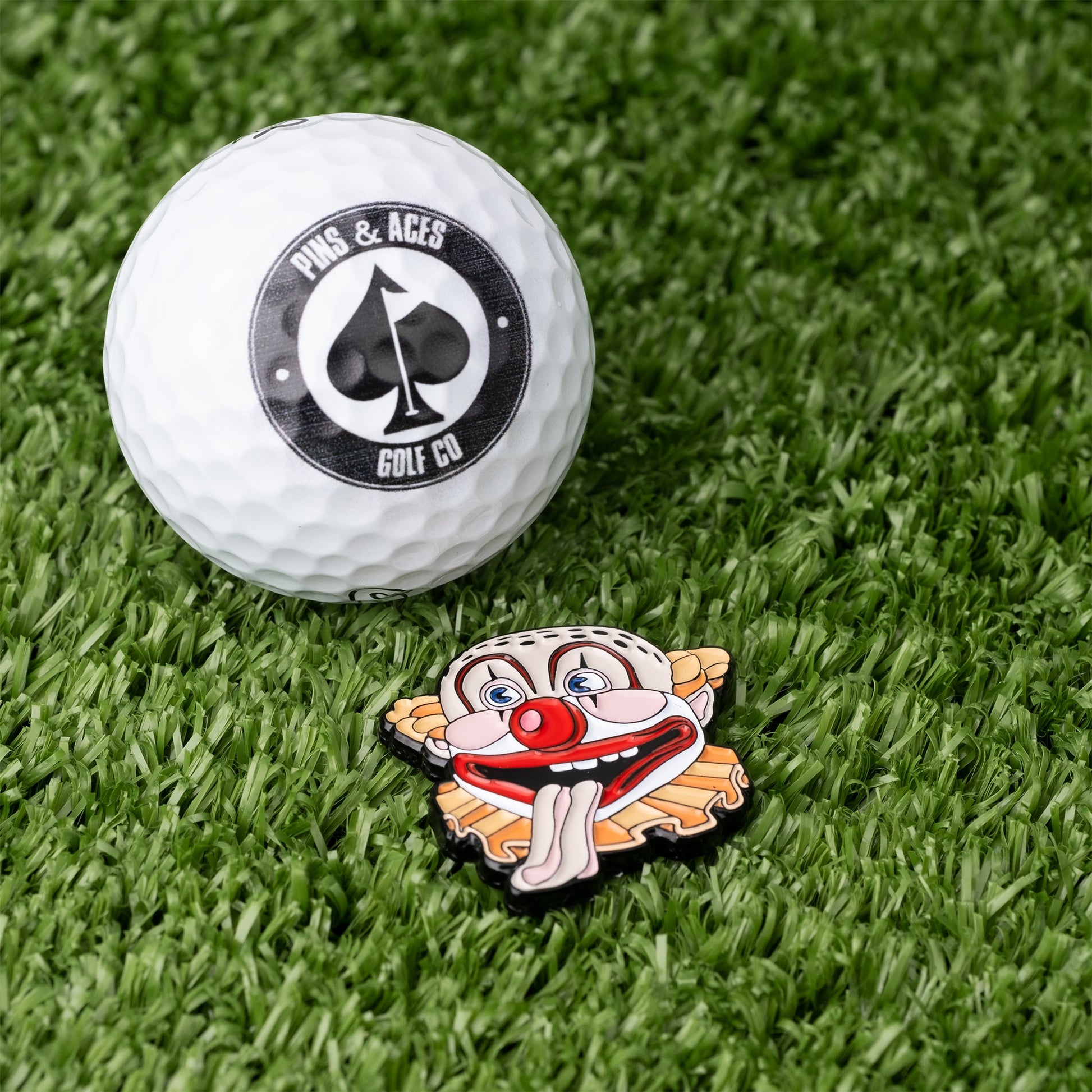 A white Pins and Aces golf ball rests on green turf beside the Clowns Mouth Ball Marker, a colorful enamel marker shaped like a cartoon clown with a red nose, tongue out, and wide eyes.