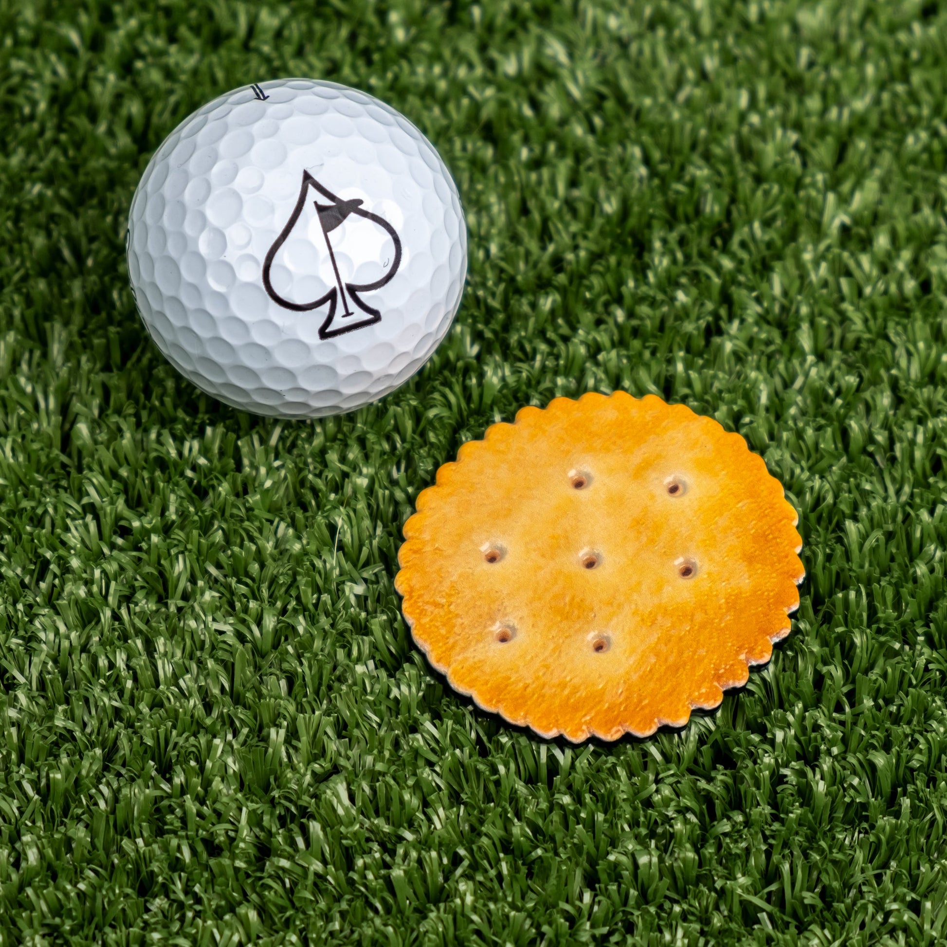 A Pins and Aces golf ball with a spade symbol rests next to The Gilmore Cracker Ball Marker, a round, golden cracker-style magnetic marker, on green artificial grass.
