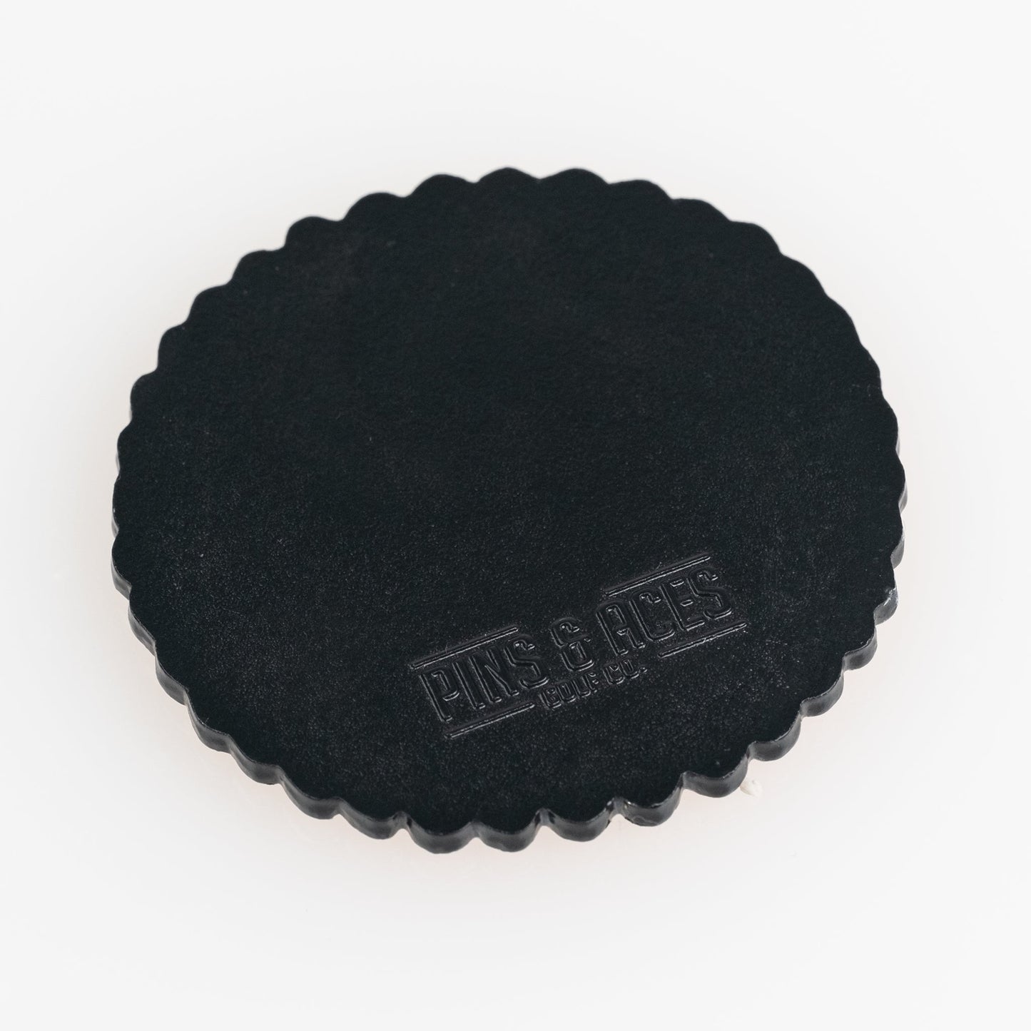 The Gilmore Cracker Ball Marker by Pins and Aces is a round, black, scalloped-edge golf ball marker with "PINS & ACES" embossed on the surface, shown on a white background.