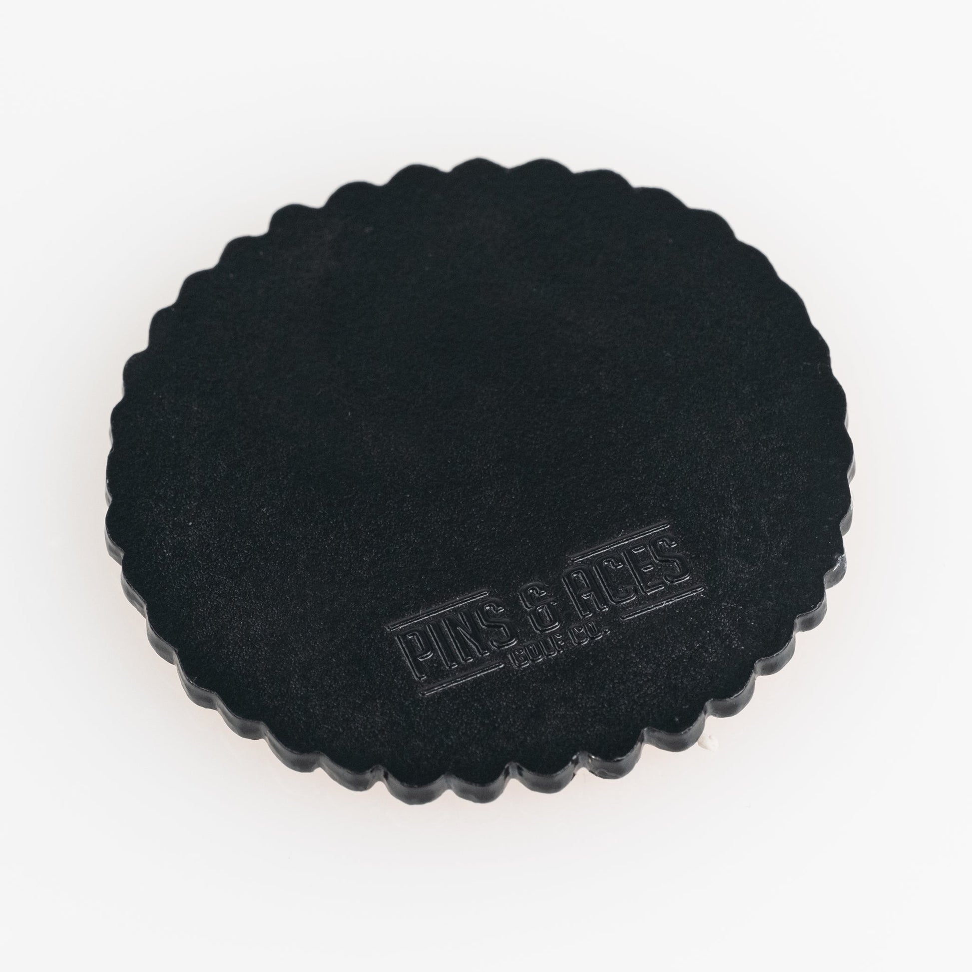 The Gilmore Cracker Ball Marker by Pins and Aces is a round, black, scalloped-edge golf ball marker with "PINS & ACES" embossed on the surface, shown on a white background.