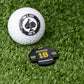 A golf ball with the Pins and Aces logo sits on grass beside a black and yellow Gilmore Jersey Ball Marker, shaped like a jersey and labeled GILMORE 18.