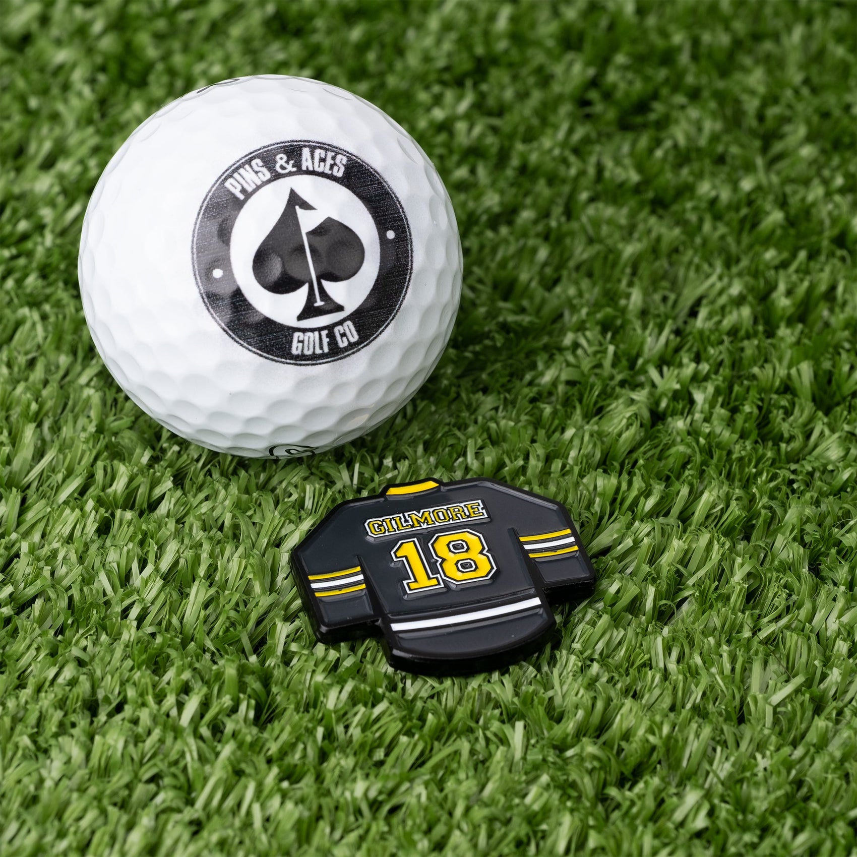A golf ball with the Pins and Aces logo sits on grass beside a black and yellow Gilmore Jersey Ball Marker, shaped like a jersey and labeled GILMORE 18.