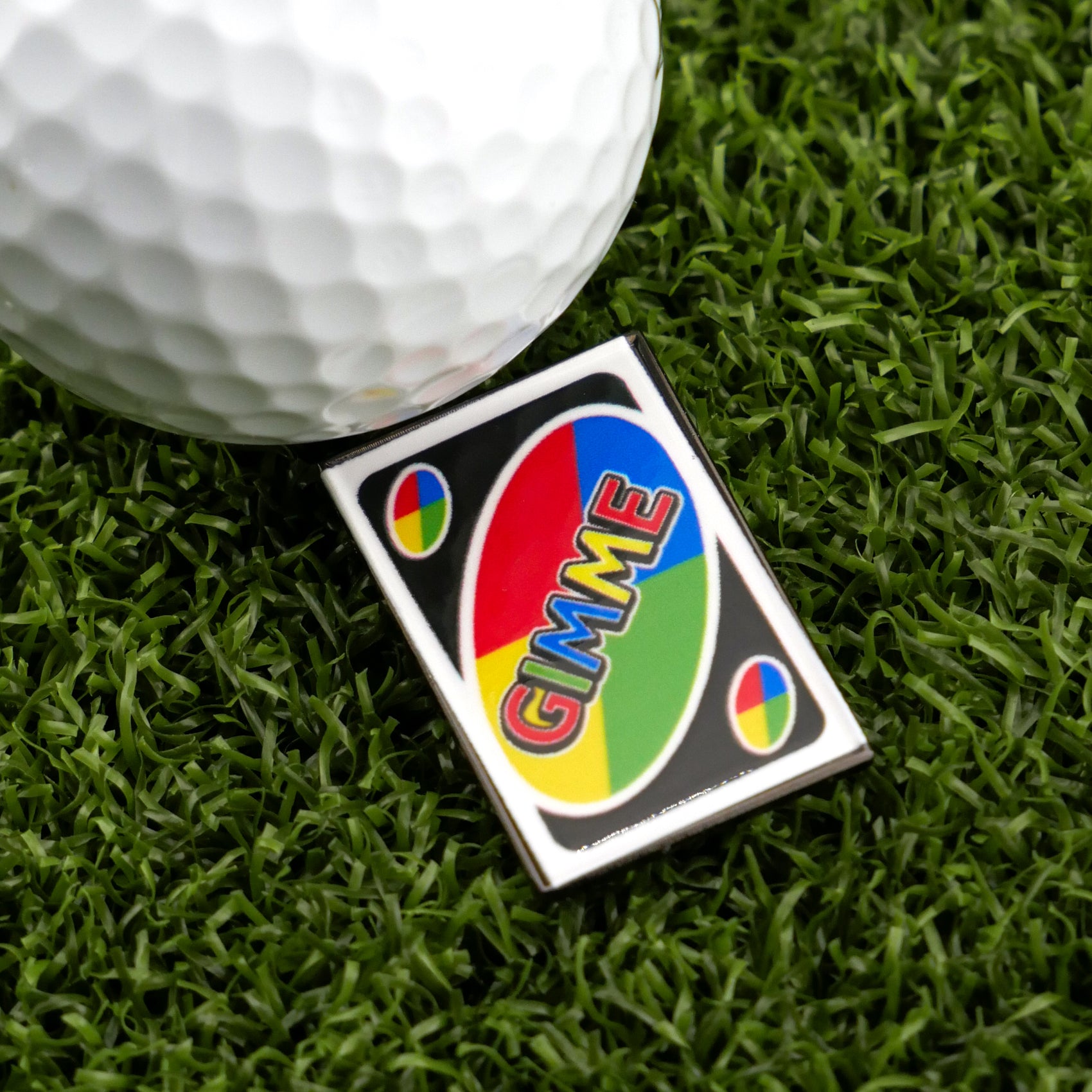 A golf ball on green grass sits next to the Gimme Wild Card by Ballmarkers.com, a premium ball marker styled like an UNO card with “GIMME” in colorful letters on a black background featuring red, blue, yellow, and green sections.