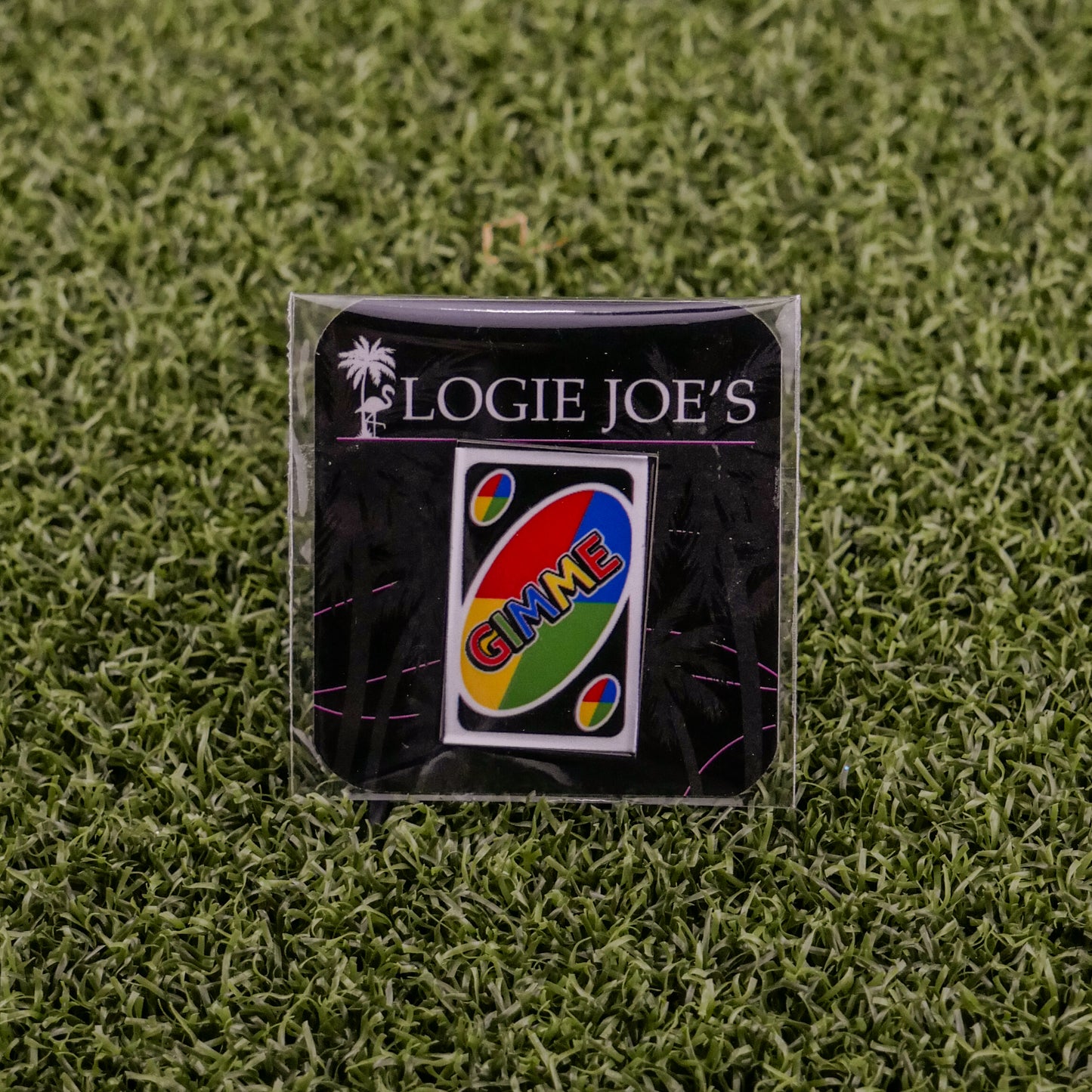 The Gimme Wild Card by Ballmarkers from Logie Joes, a premium UNO-inspired ball marker with GIMME, is showcased on artificial green grass and attached to a branded LOGIE JOES card.