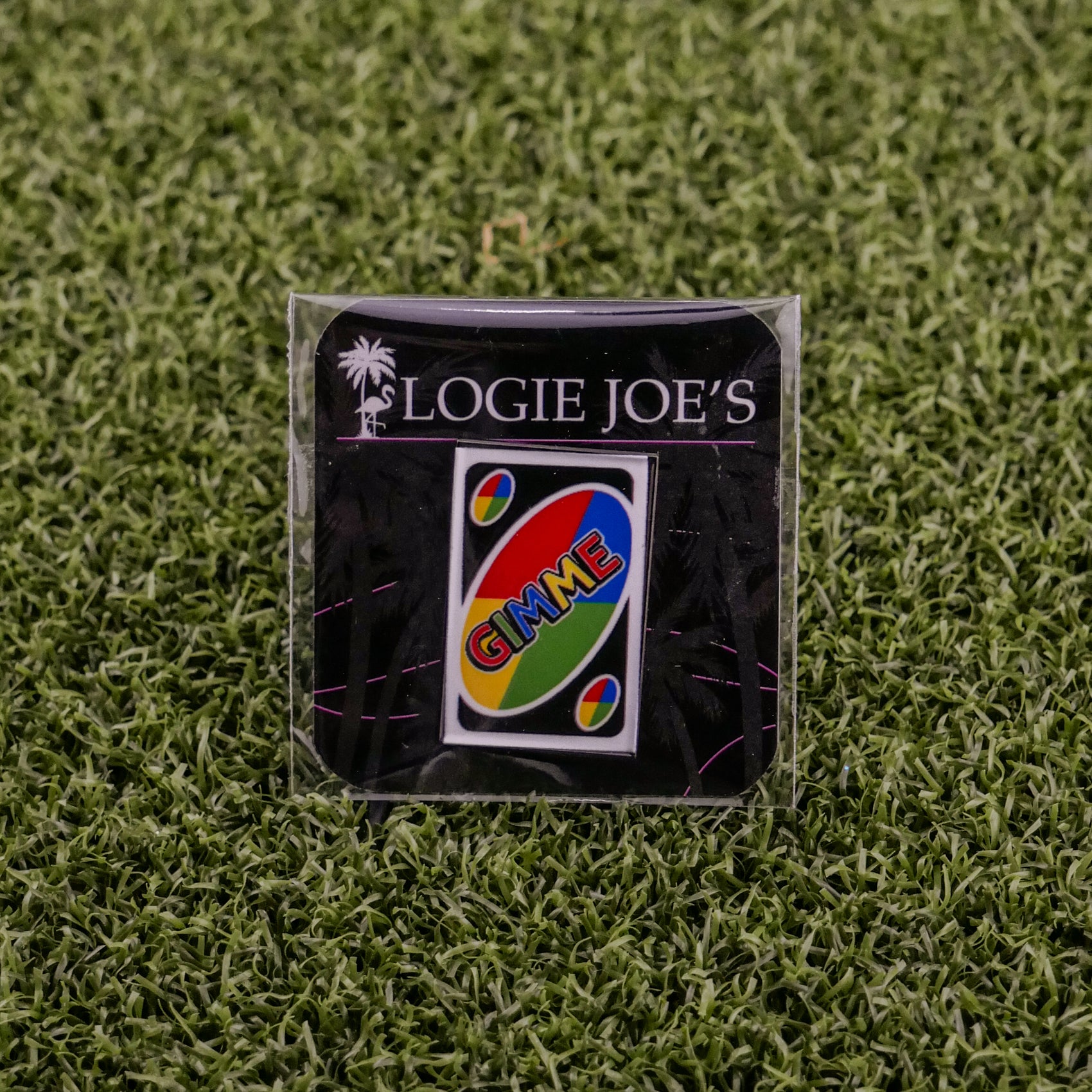 The Gimme Wild Card by Ballmarkers from Logie Joes, a premium UNO-inspired ball marker with GIMME, is showcased on artificial green grass and attached to a branded LOGIE JOES card.