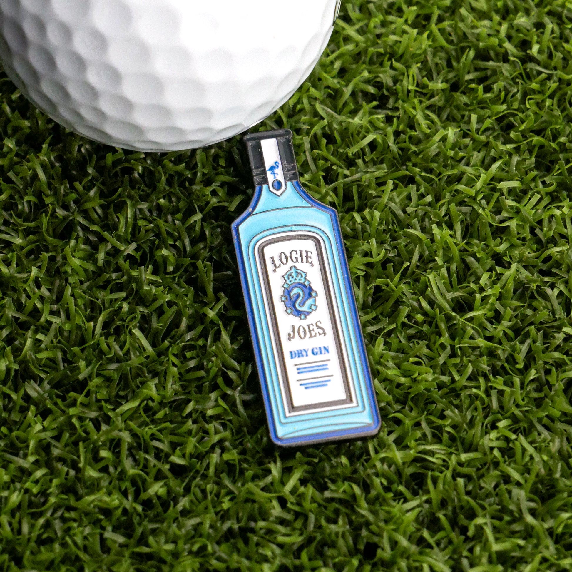A bottle-shaped Gin Ball Marker from Logie Joe's Golf rests on green artificial grass beside a white golf ball, making it a unique golf accessory and the perfect gift for golfers.
