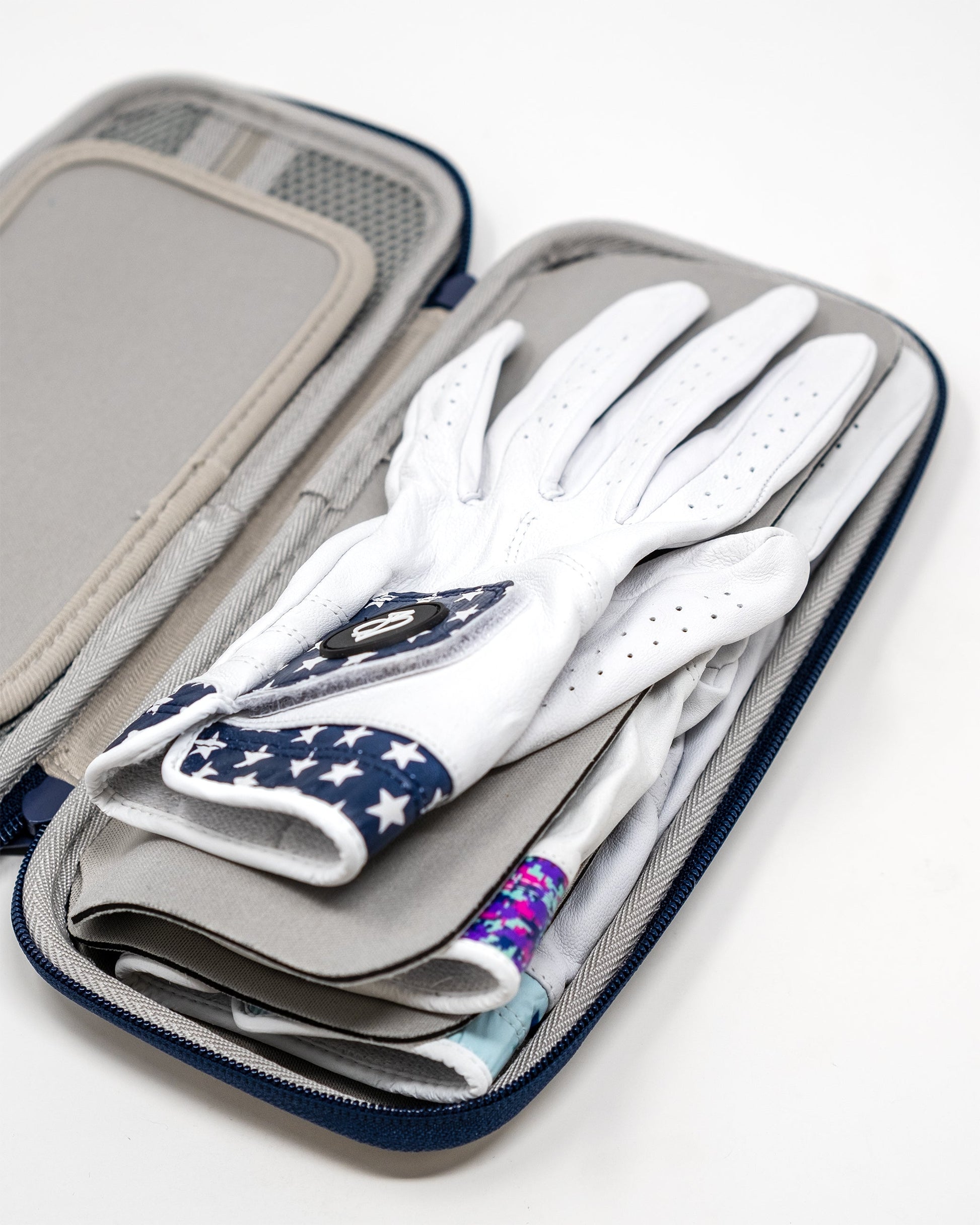 A close-up of the Pins and Aces Glove & Accessory Caddie - Navy shows it open with neatly stacked golf gloves, including a white glove with a blue star-patterned strap, displayed on a white background.