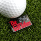 A close-up of a golf ball beside Logie Joe's Golf "The Golf Father" black marker pin on grass, featuring red and white text styled like The Godfather logo with a puppet hand—an amusing golf accessory for fans.