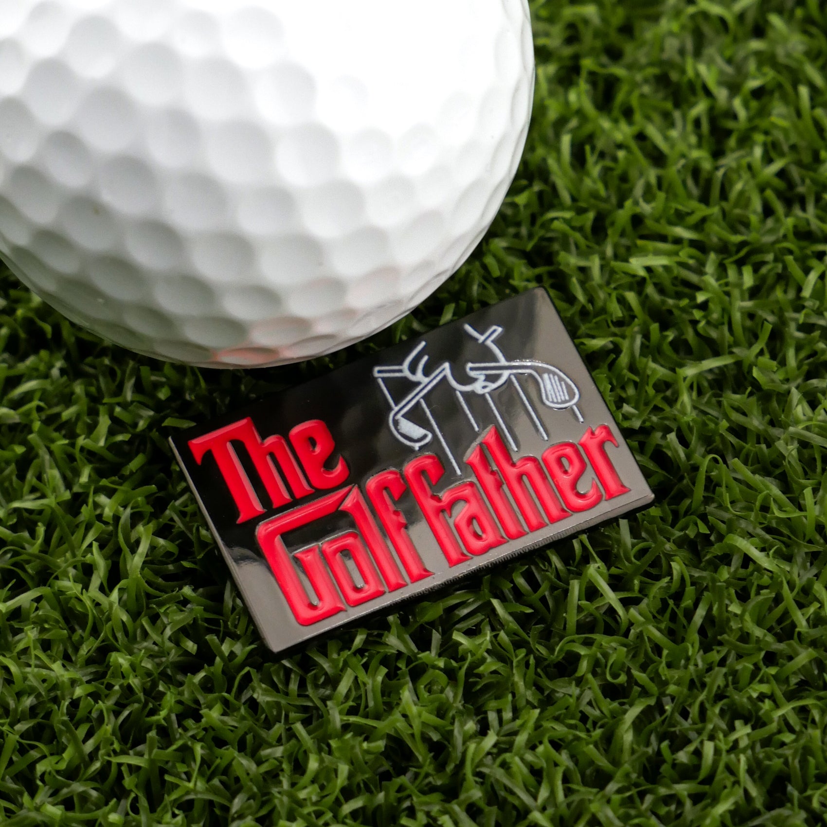 A close-up of a golf ball beside Logie Joe's Golf "The Golf Father" black marker pin on grass, featuring red and white text styled like The Godfather logo with a puppet hand—an amusing golf accessory for fans.