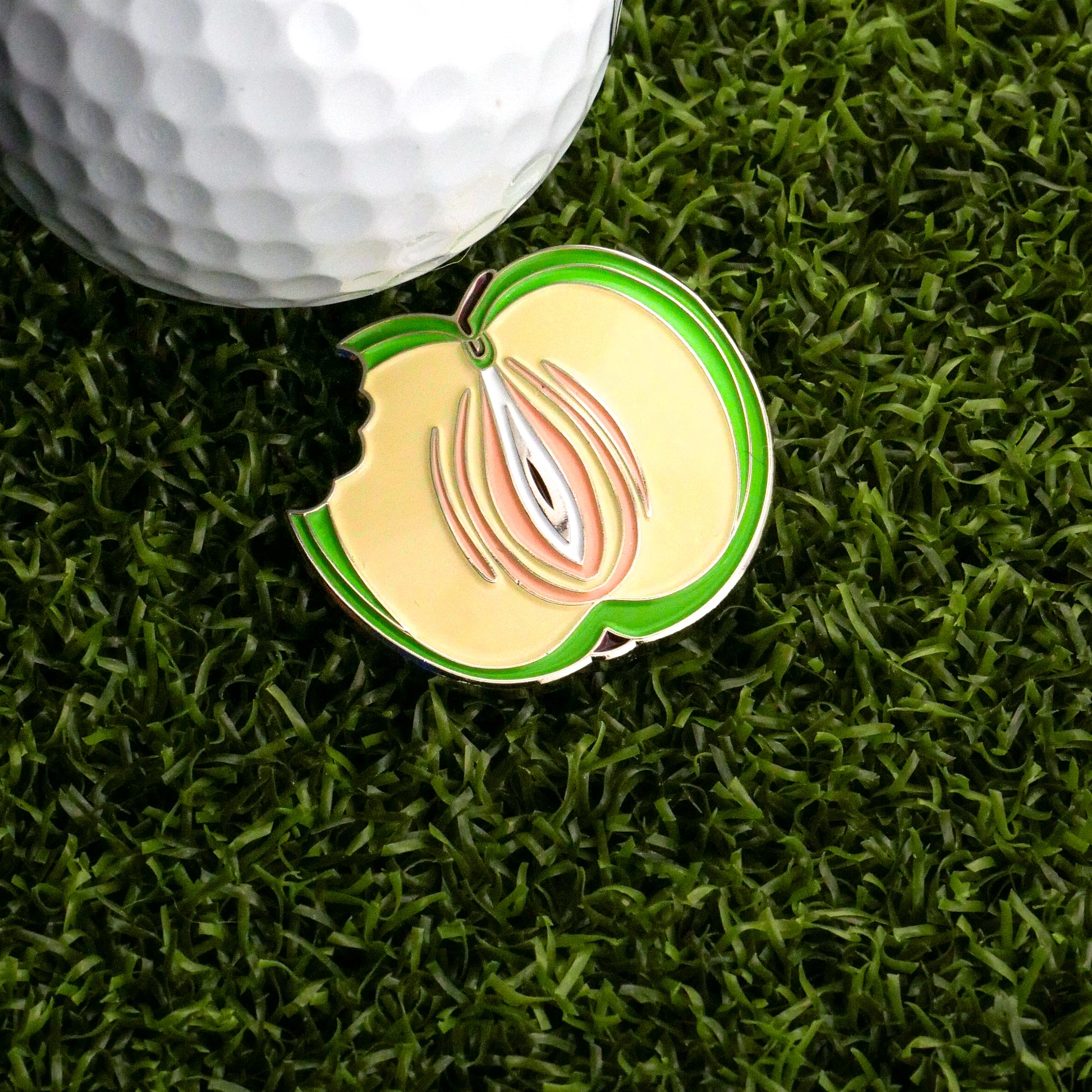 A golf ball sits on artificial green grass beside the Bunkers & Rough Green Apple Vagina Ball Marker, a novelty marker shaped like a bitten apple half with seeds and a green outline.