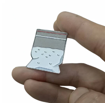 A hand holds the Ground Score Ball Marker by Golf Gear Heaven, a clever enamel pin shaped like a clear plastic bag with a red seal and white granules, designed as a unique golf accessory.