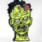 The Mutant Zombie - Ball Marker by Pins and Aces features a cartoonish green zombie head with exposed brains, black messy hair, and an angry look, displayed on a plain white background.