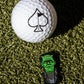 A Pins and Aces Frankenstein Ball Marker and a white golf ball with a spade symbol sit on green turf beside a small Frankenstein’s monster figurine—a unique, premium accessory for golfers seeking standout style.