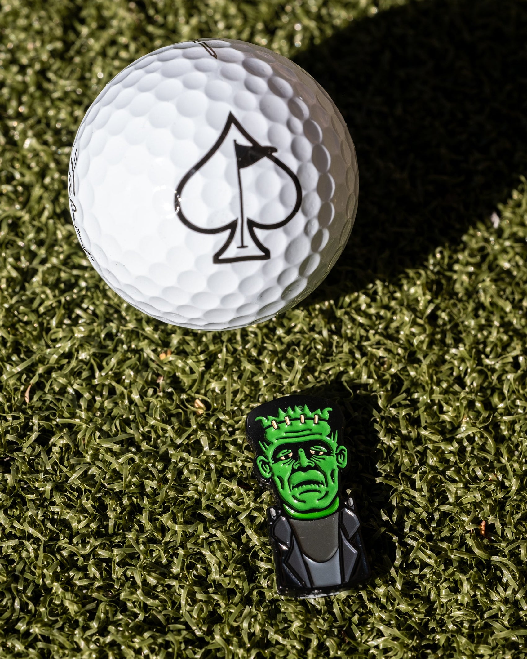 A Pins and Aces Frankenstein Ball Marker and a white golf ball with a spade symbol sit on green turf beside a small Frankenstein’s monster figurine—a unique, premium accessory for golfers seeking standout style.