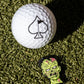 A white golf ball with a black spade symbol sits on artificial grass beside the Pins and Aces Mutant Zombie - Ball Marker, featuring a colorful green cartoon zombie face patch.