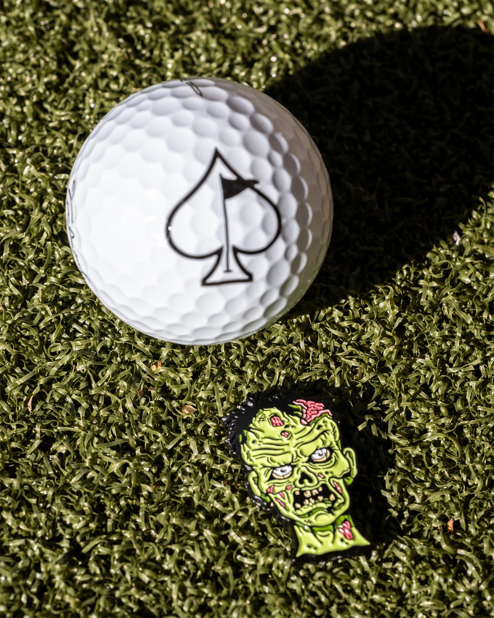A white golf ball with a black spade symbol sits on artificial grass beside the Pins and Aces Mutant Zombie - Ball Marker, featuring a colorful green cartoon zombie face patch.