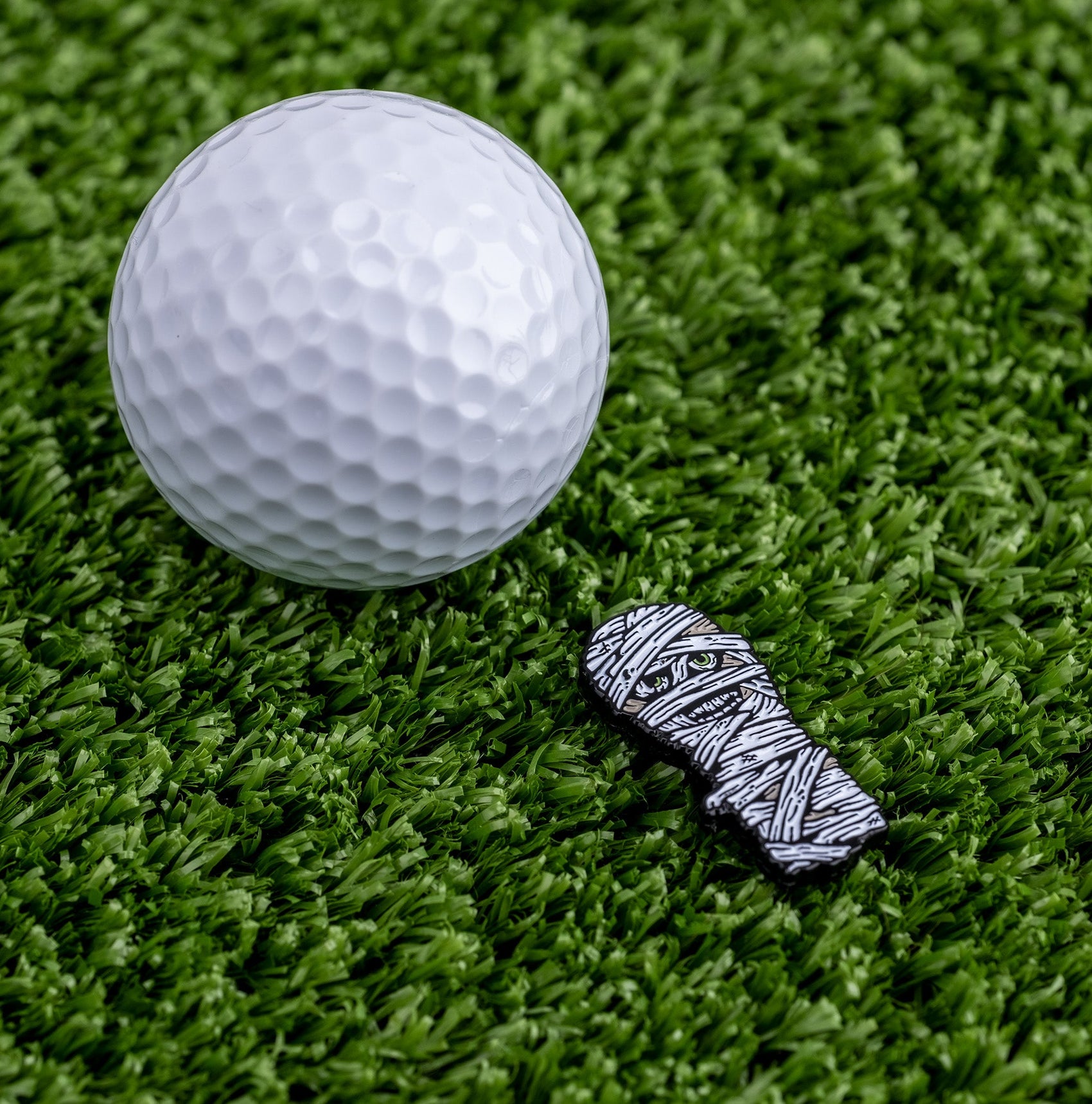 A white golf ball and the Pins and Aces Mummy Ball Marker, a small enamel-finished magnetic marker, sit on green artificial turf.