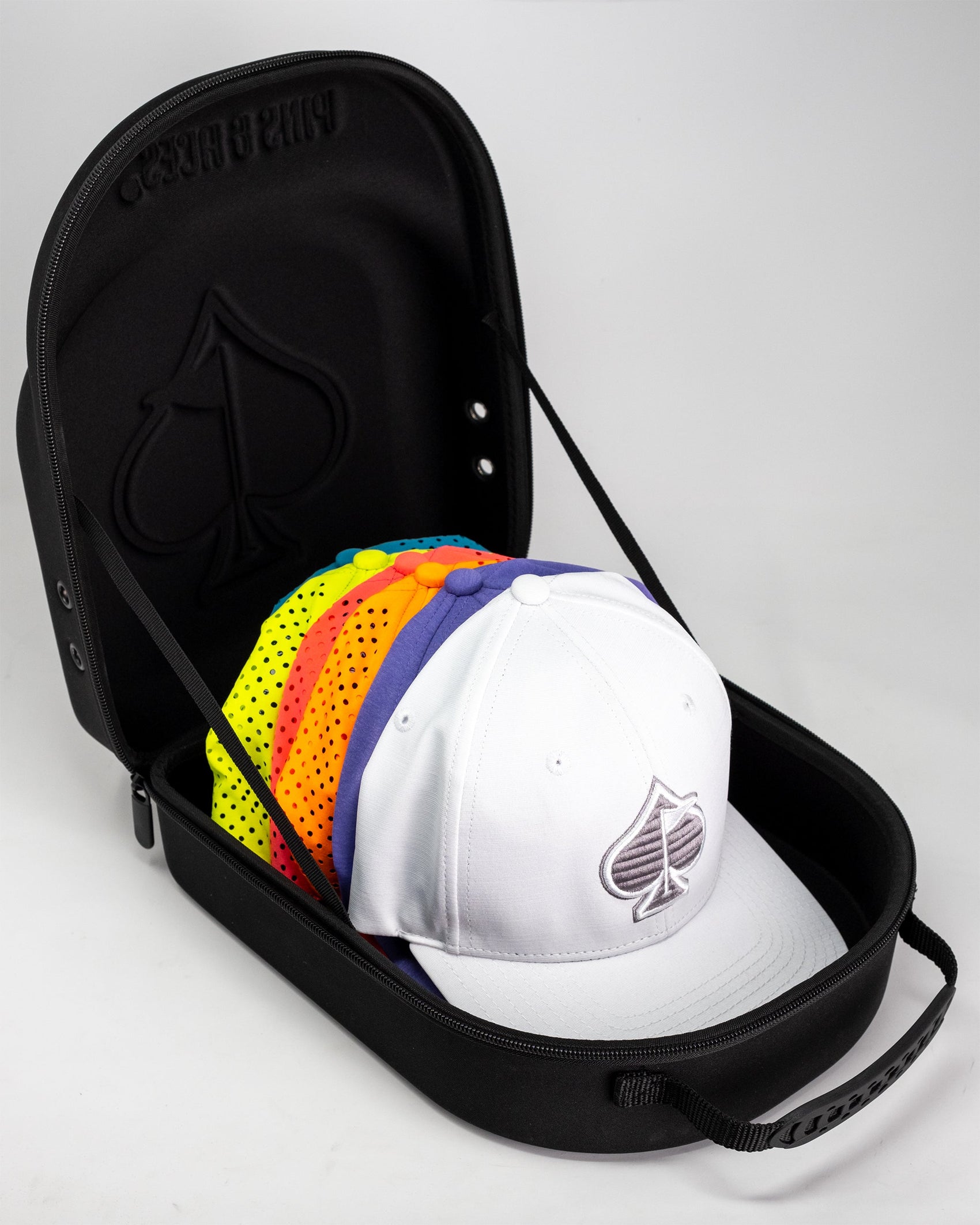The Pins and Aces Hat Caddie, shown partially open in black, holds several colorful baseball caps, with a white cap featuring a spade logo on top; its interior displays a spade-shaped imprint.