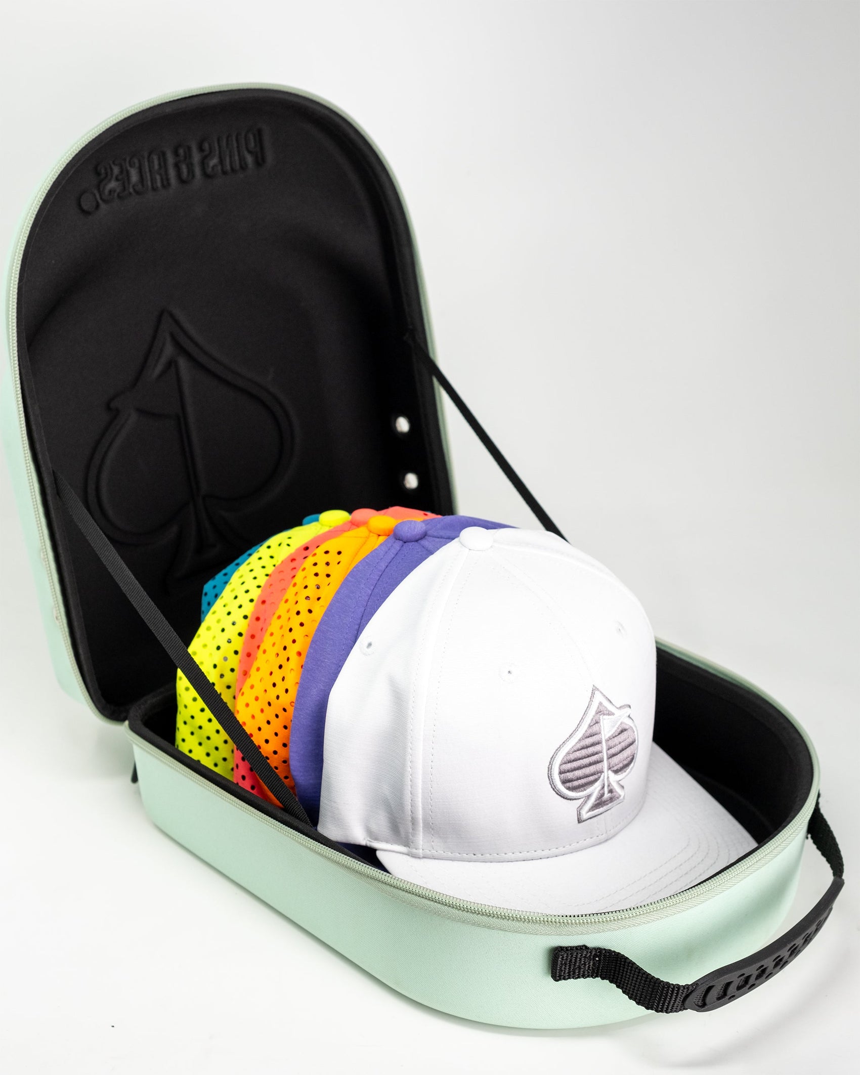 The Pins and Aces Hat Caddie, shown open on a white background, is a mint green case embossed with the brand’s name, neatly storing several colorful caps with a white spade logo hat on top.