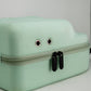 The Pins and Aces Hat Caddie is a light green golf hat case with a zippered closure and two metal grommets, resembling eyes and a mouth, shown against a plain light background.