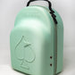 The Hat Caddie by Pins and Aces is a light green, semi-hard golf hat case with a handle, front spade embossing, "PINS & ACES" above the spade, zipper closure, and two metal ventilation holes on the side.