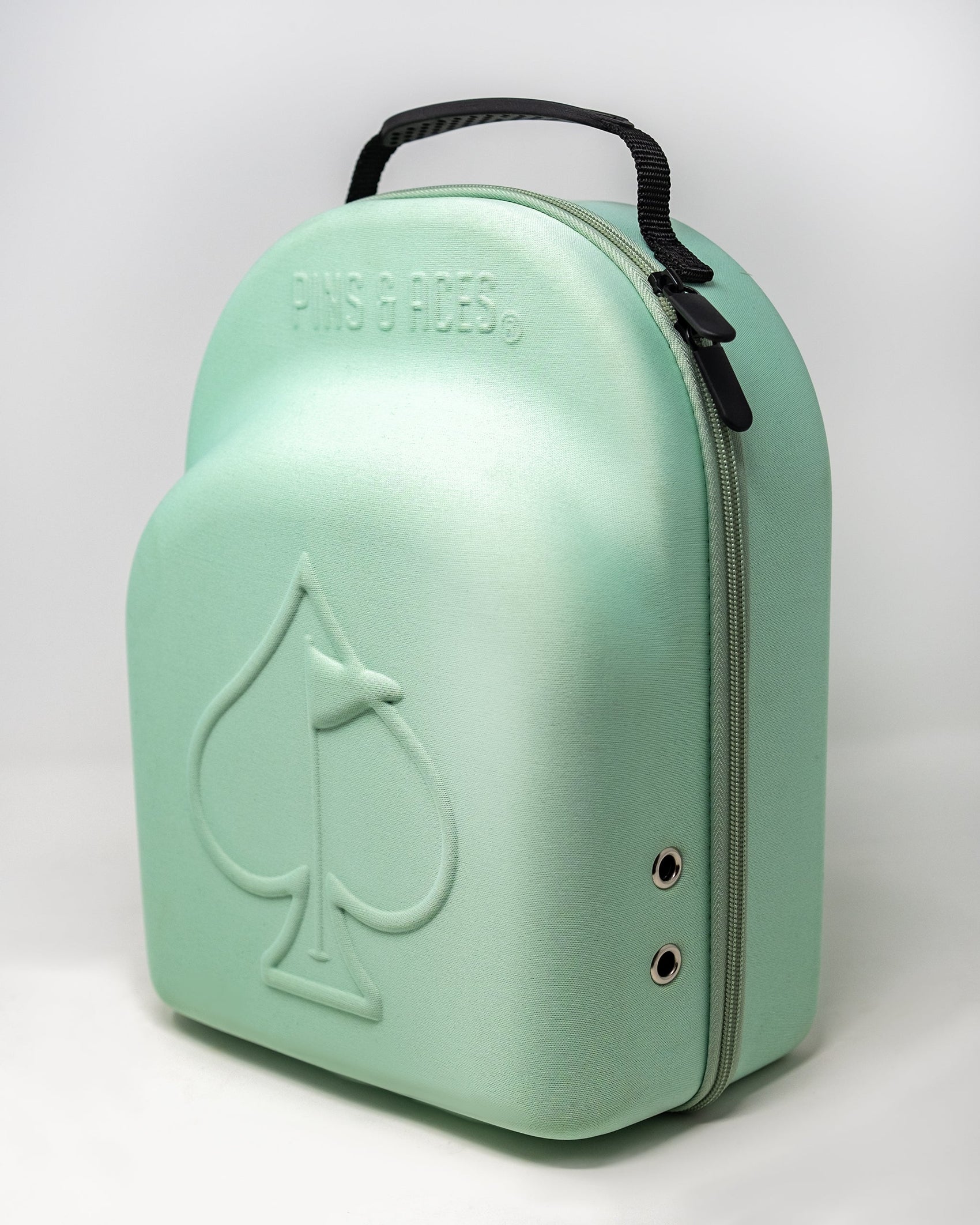 The Hat Caddie by Pins and Aces is a light green, semi-hard golf hat case with a handle, front spade embossing, "PINS & ACES" above the spade, zipper closure, and two metal ventilation holes on the side.