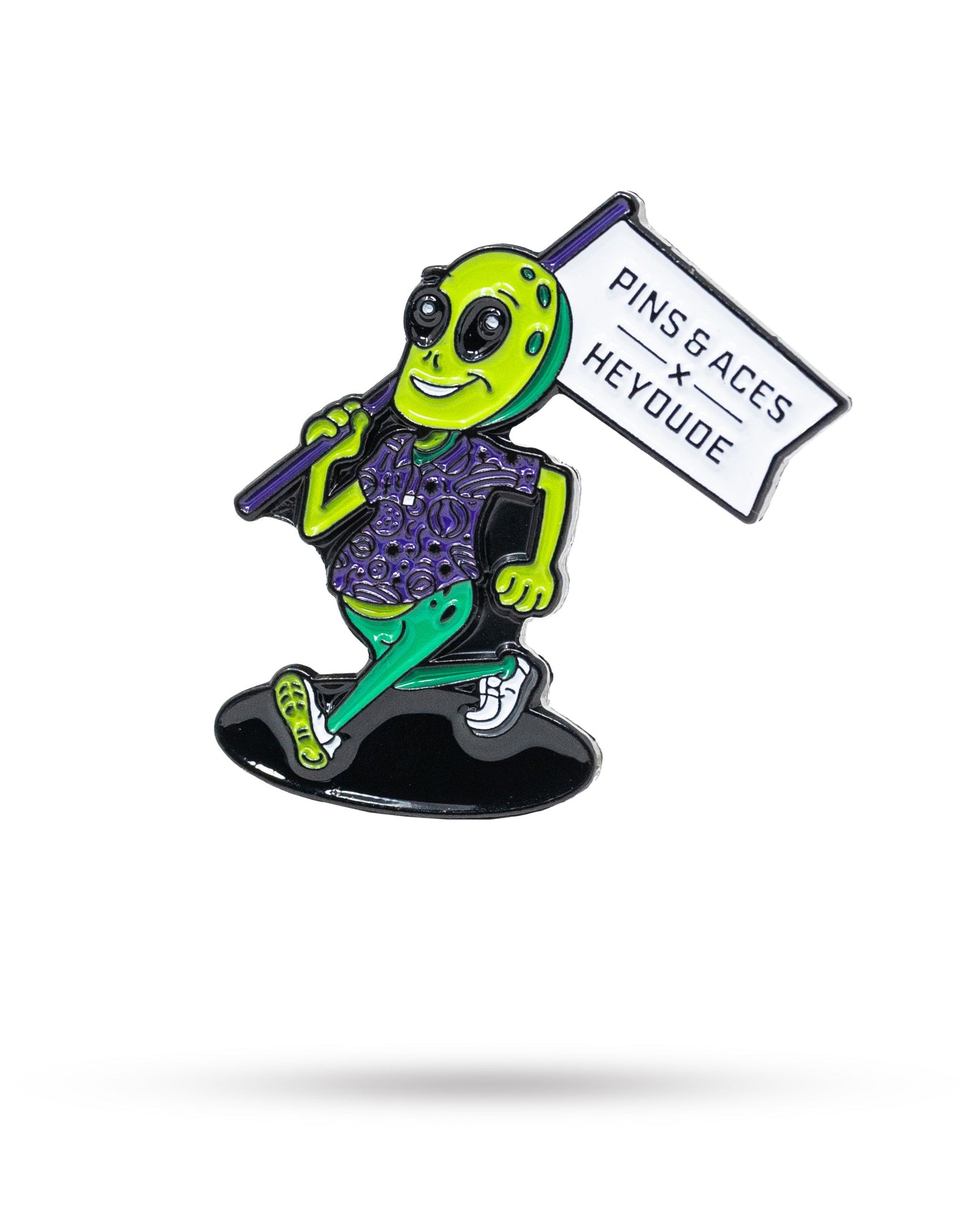 A cartoon alien in a purple patterned shirt and green pants walks while holding a PINS AND ACES flag, showing off the HEYDUDE – Alien Ball Marker by Pins and Aces, displayed on a white background.