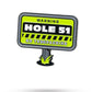 A neon yellow sign reads "WARNING HOLE 51 NO TRESPASSING" in bold black and white letters with grass graphics at the bottom, displayed beside the HEYDUDE - Hole 51 Ball Marker by Pins and Aces.
