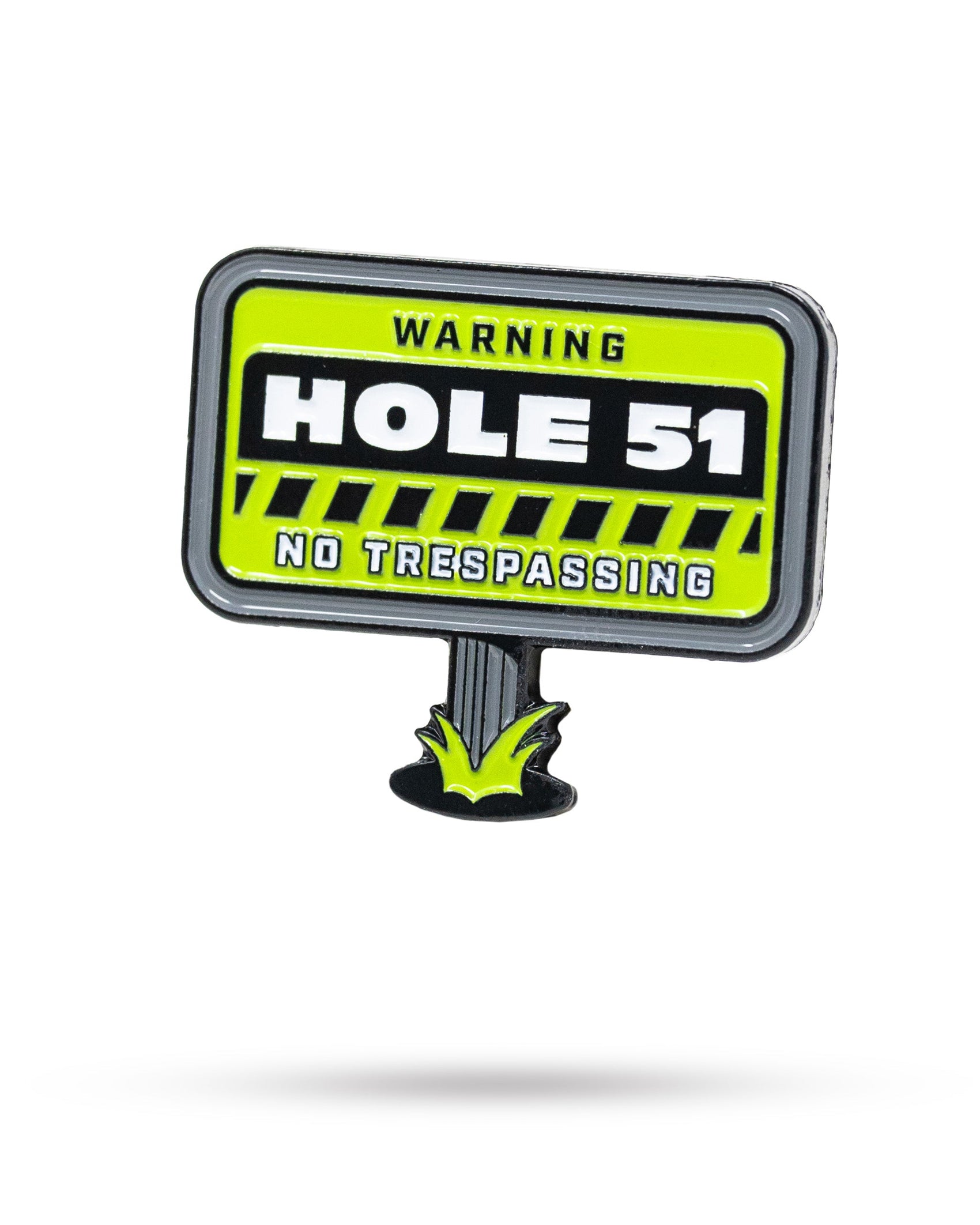 A neon yellow sign reads "WARNING HOLE 51 NO TRESPASSING" in bold black and white letters with grass graphics at the bottom, displayed beside the HEYDUDE - Hole 51 Ball Marker by Pins and Aces.