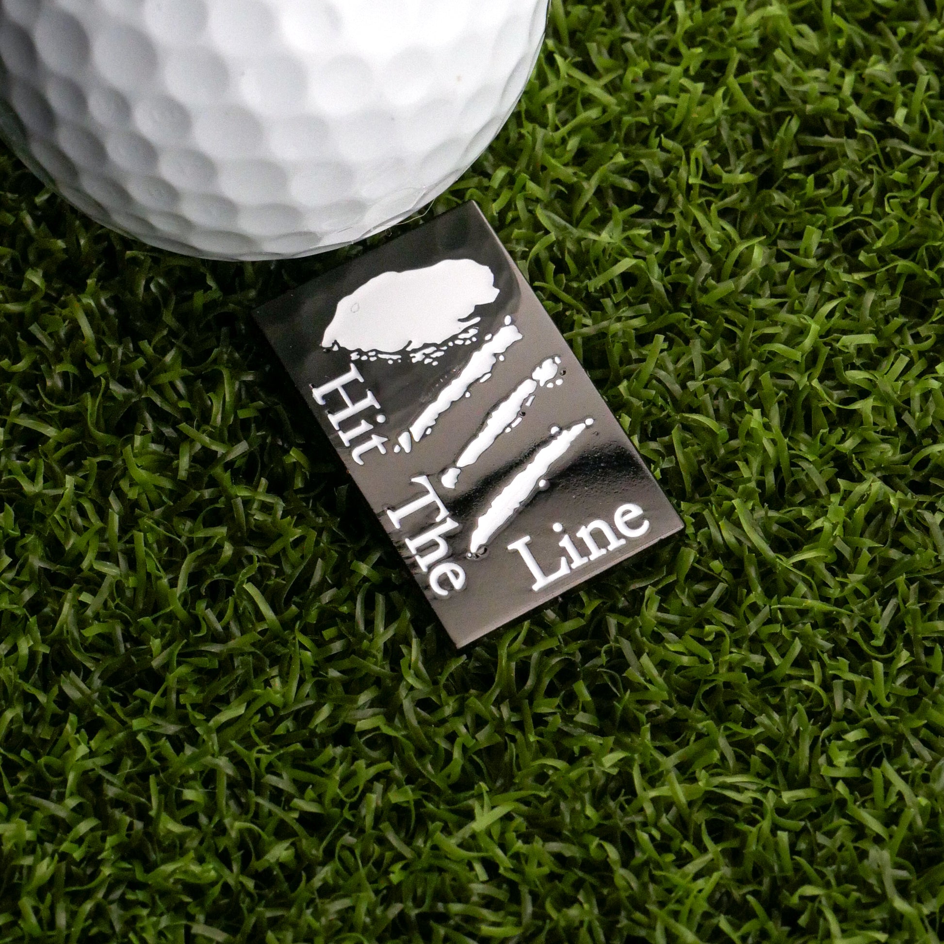 A close-up of a golf ball on artificial grass beside the Logie Joe's Golf Hit the Line Ball Marker, a rectangular black marker with white "Hit The Line" text, claw-like scratch marks, and a splash graphic above the lettering.